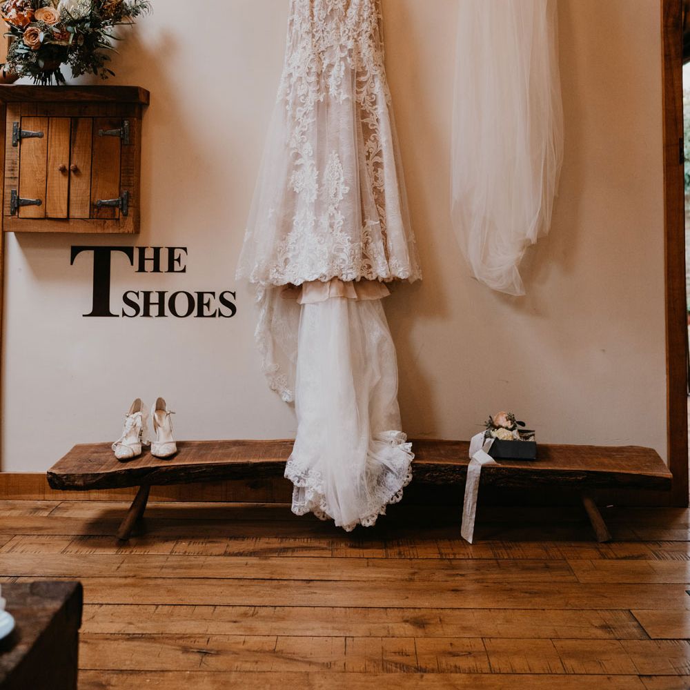 Lace wedding dress with train hands from wooden beam next to veil, shoes and bouquet at the Tythe Barn 