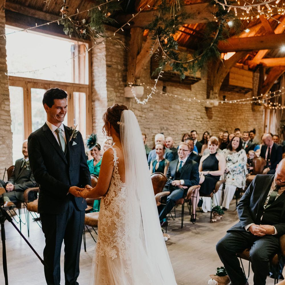 Bride in open back lace wedding dress and veil stands with groom in dark suit and blue tie during rustic wedding ceremony