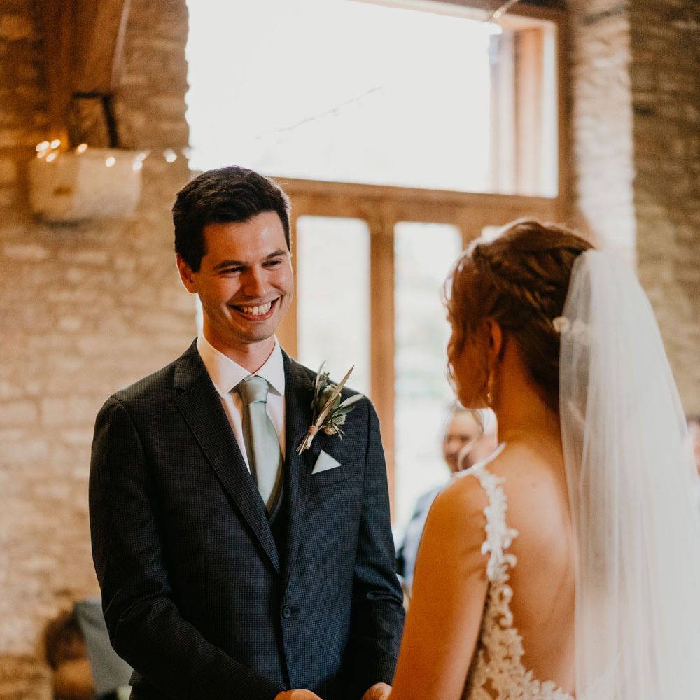 Smiling groom in dark suit, light blue tie and pocket square stands holding hands with bride in lace wedding dress during wedding ceremony