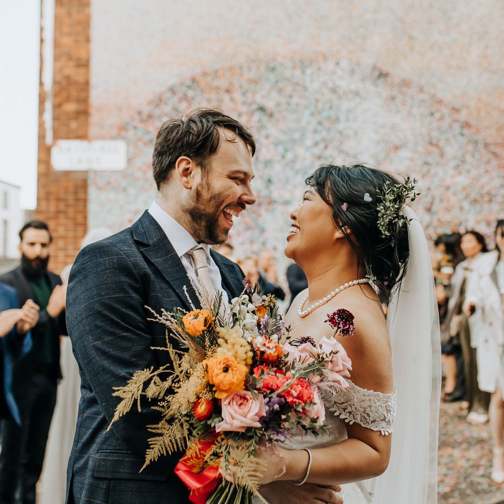 Smiling bride in pearl necklace and veil holding wild mixed wedding bouquet stands whipping groom in blue check suit with light pink tie after confetti toss