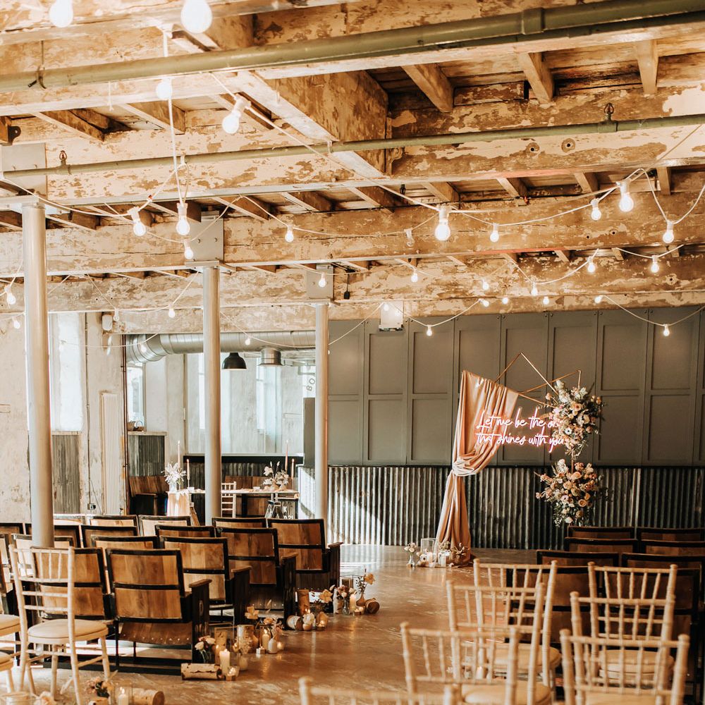 Interior of industrial boho wedding ceremony with pastel florals, bulb string lights and wooden chairs