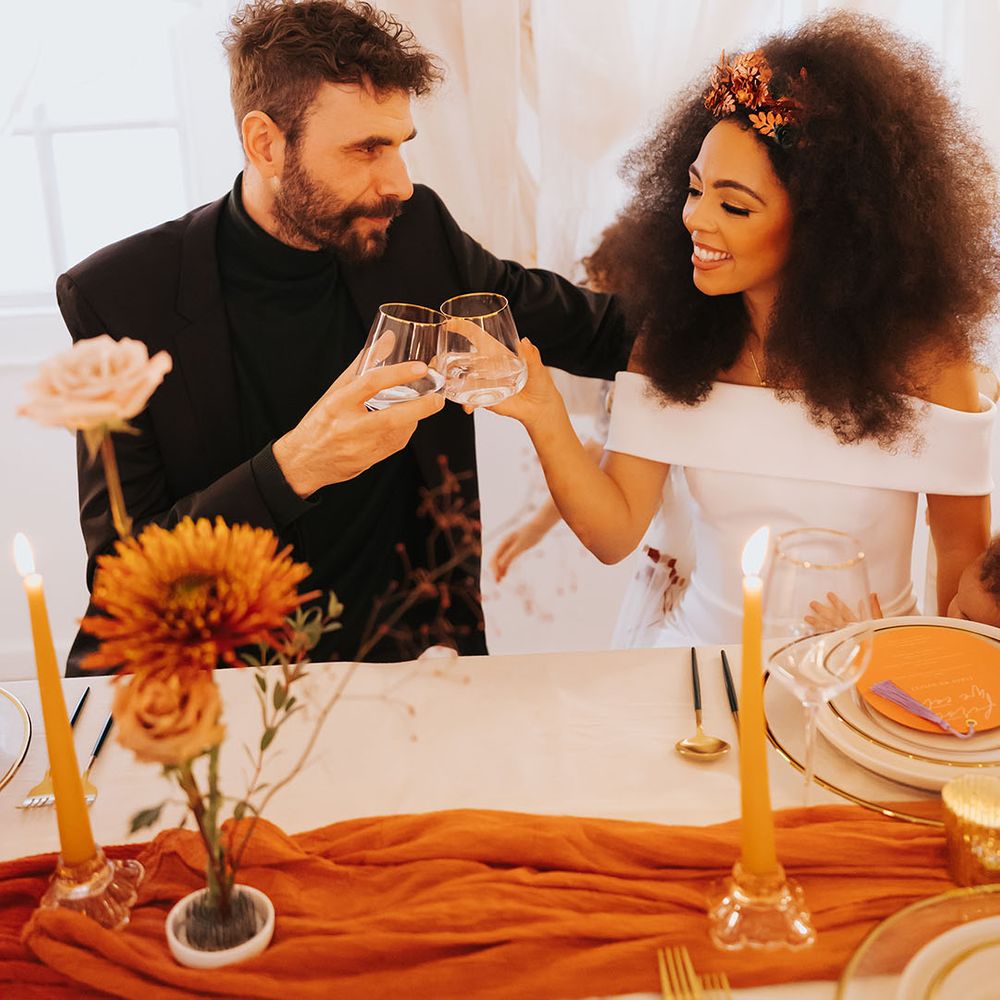 Black bride in an off the shoulder wedding dress with afro hair and hair accessory clinking glasses with her groom in a black suit 