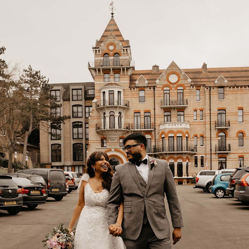 Bride in white traditional wedding dress and mixed flower neutral bouquet with groom in grey suit and black bow tie walking at The Petersham Hotel in Richmond