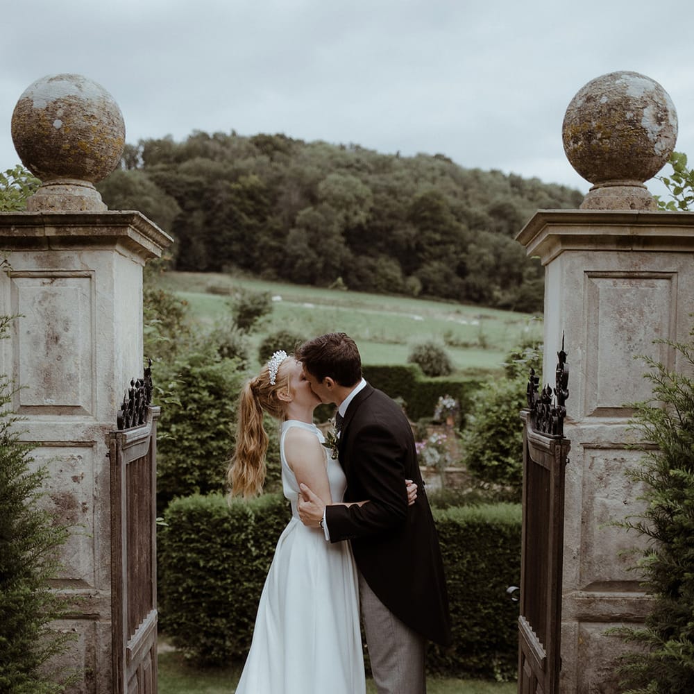 Groom in morning suit with grey trousers kisses bride in the gardens at Owlpen Manor 