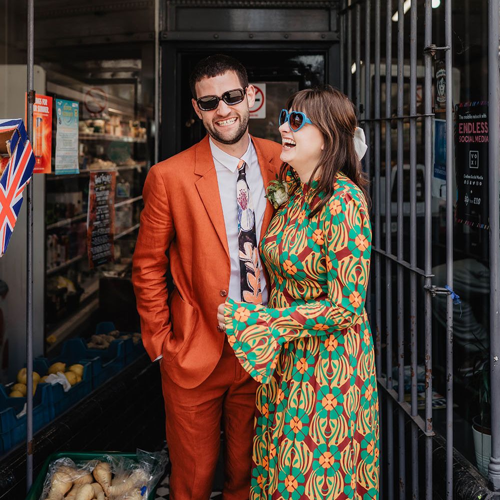 Bride wears blue heart shaped sunglasses and brightly coloured wedding gown next to her groom in an orange suit 