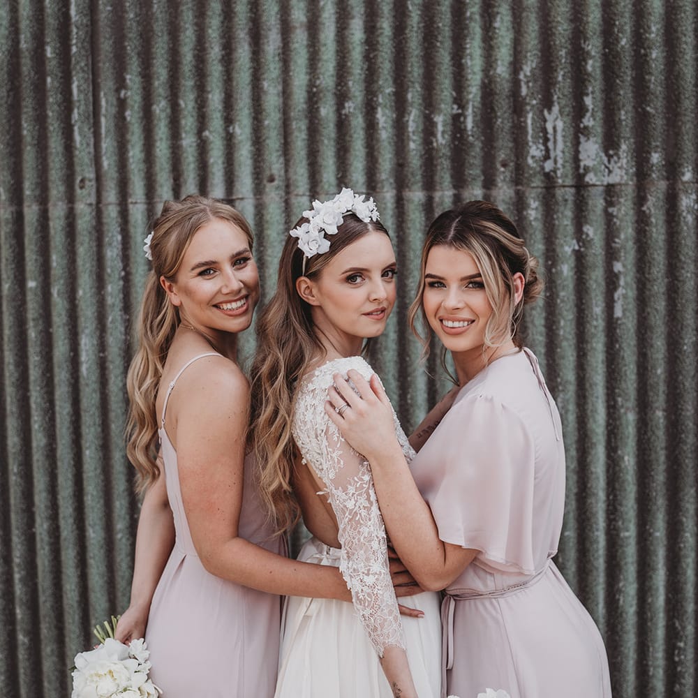 Bride wearing a flower headband in a lace wedding dress poses with the bridesmaids in pale pink dresses