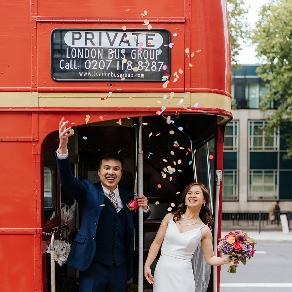 Groom throws colourful confetti into the air beside his bride on red London bus 