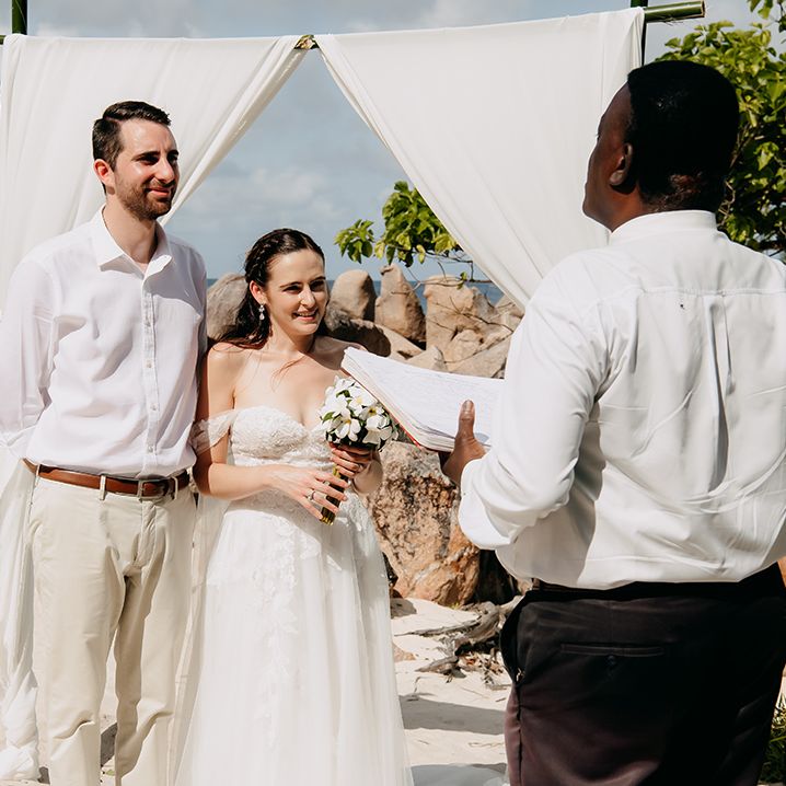 Bride in off the shoulder lace wedding dress with sweetheart neckline and lace floral details and cathedral length veil holding white tropical bouquet and  groom in crisp white shirt, light coloured chinos, tan leather belt and white trainers eloping in Seychelles
