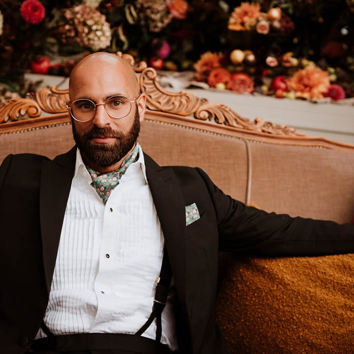 Groom on vintage orange loveseat wearing classic black tuxedo and black suspenders with hand painted silk cravat and handkerchief 