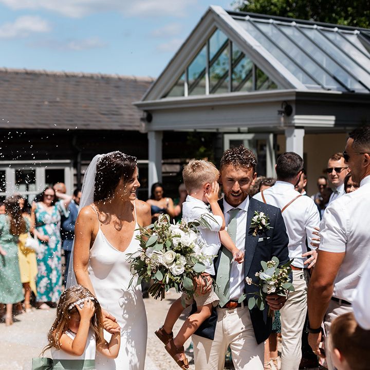 Bride in silk Grace Loves Lace wedding dress leaves wedding ceremony with her groom and children as white confetti is throw around them