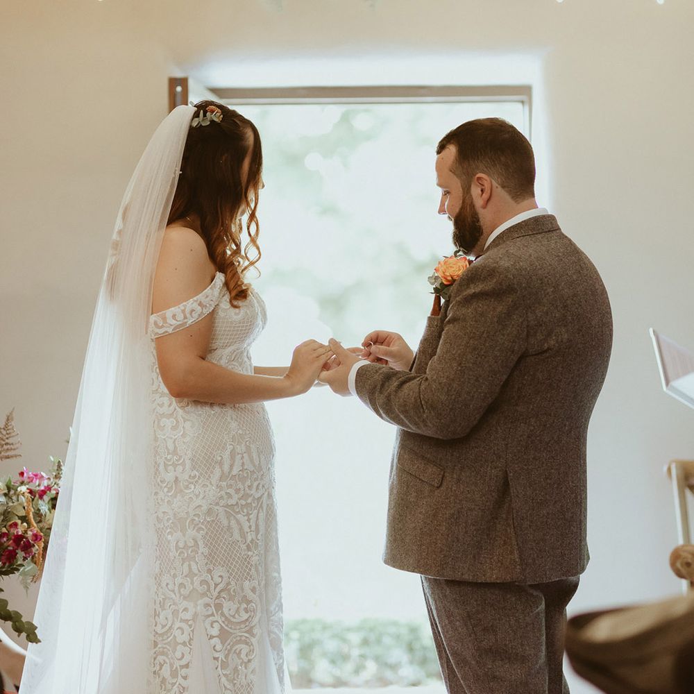 Groom in three piece tweed suit and orange buttonhole puts the ring on the bride's finger wearing a boho fitted off the shoulder lace wedding dress