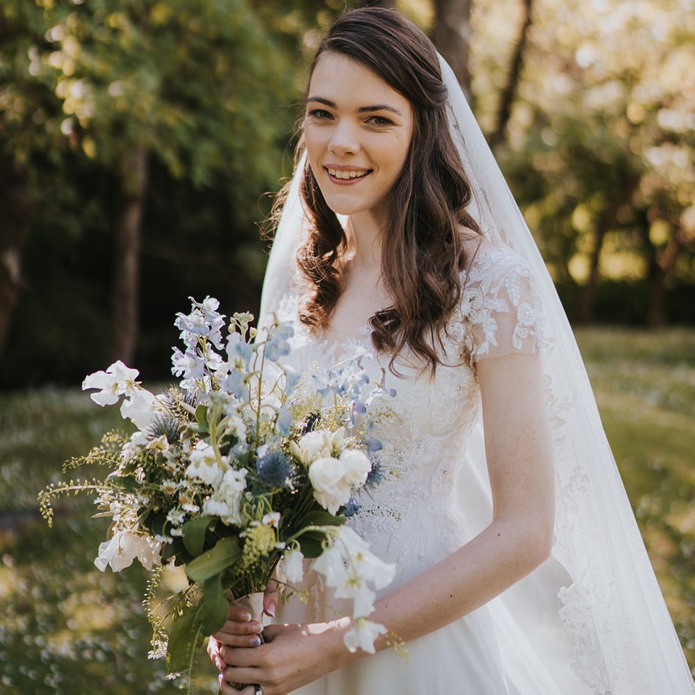 Bride in short sleeve lace wedding dress with veil holding white and blue bouquet of flowers for the wedding 