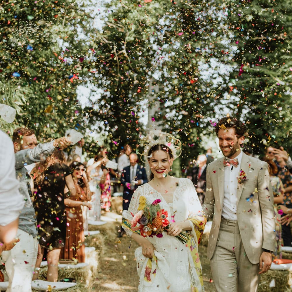 Confetti exit with bride in lace wedding dress and bridal crown carrying bright pink bridal bouquet with groom in greige suit and colourful bow tie 