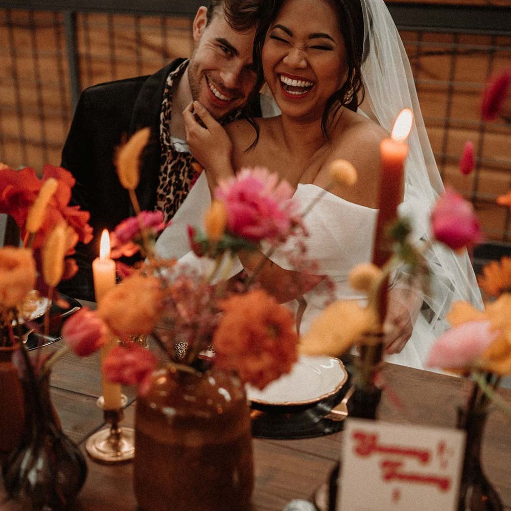 Bride in off shoulder tulle short wedding dress with church length veil sitting with groom in wet look black grooms suit with leopard print shirt and pink and orange boutonniere at rustic wedding tablescape with vinyl record place mats, gold rimmed plates, gold cutlery, colourful pink, red and orange floral centrepieces in bottles and tapered candles 