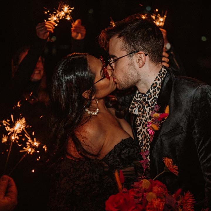 Bride in black ruffle off shoulder reception dress and red drip effect sunglasses standing with groom in black shirt and black drip effect sunglasses at German shoot with vinyl wedding arch doing fun sparkler send off