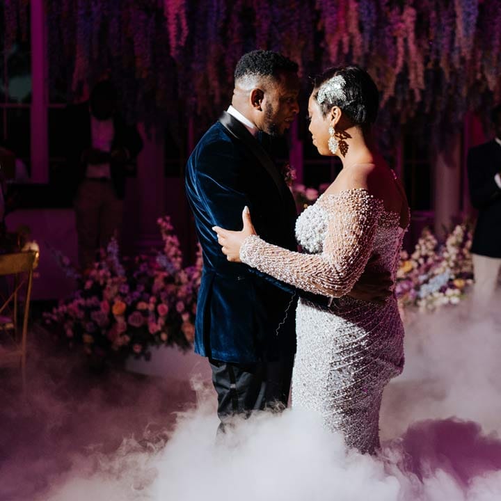 Groom in velvet midnight blue double breasted groom’s blazer, velvet bowtie and peach garden rose boutonniere dancing with bride in illusion v neck sheer off shoulder long sleeve pearl embellished wedding gown at Syon Park vow renewal 