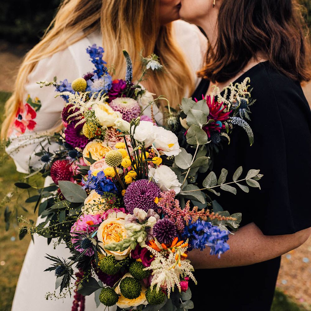 Colourful wildflower wedding bouquet with pink, purple, and yellow flowers held by two brides 
