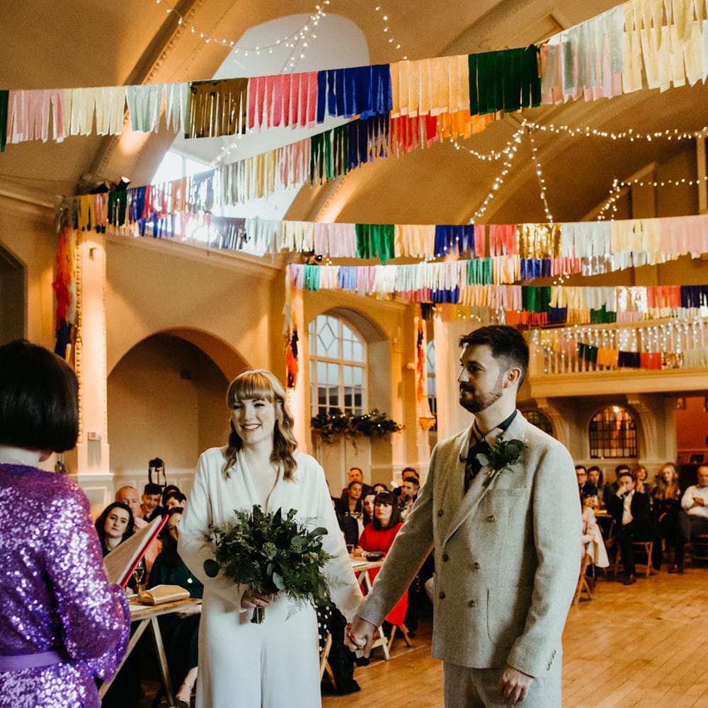 Bride in a jumpsuit holding hands with her groom in a grey double breasted suit at the altar 