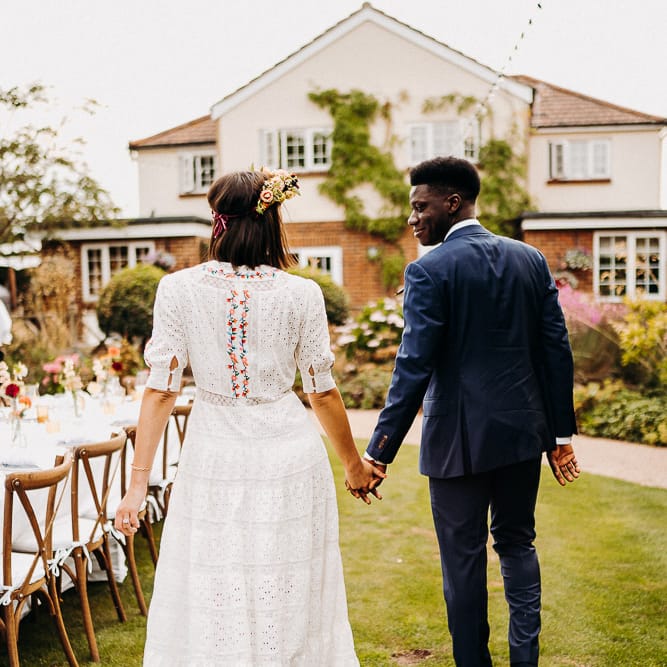 Groom in a navy blue suit holding hands with his bride in a bohemian wedding dress as they walk around the table at their outdoor reception table 