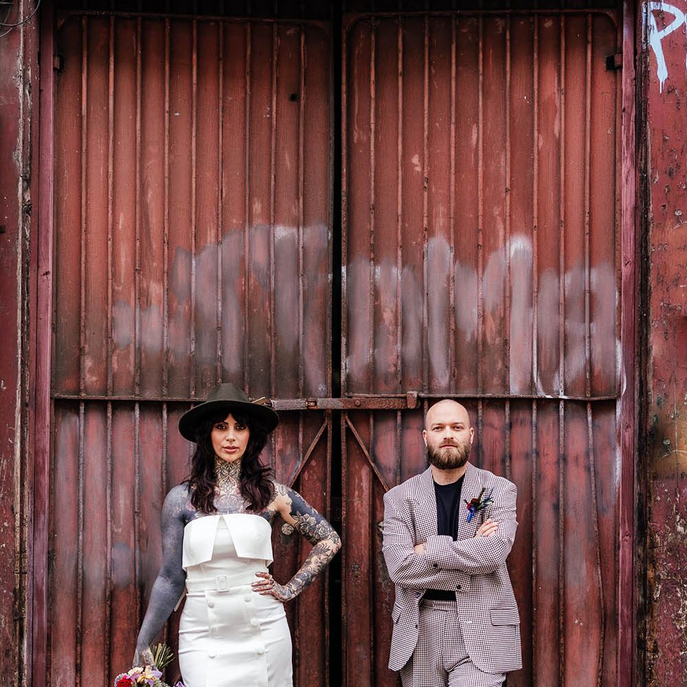 Alternative bride and groom in a strapless wedding dress and fedora hat and check suit standing neck to a corrugated iron container