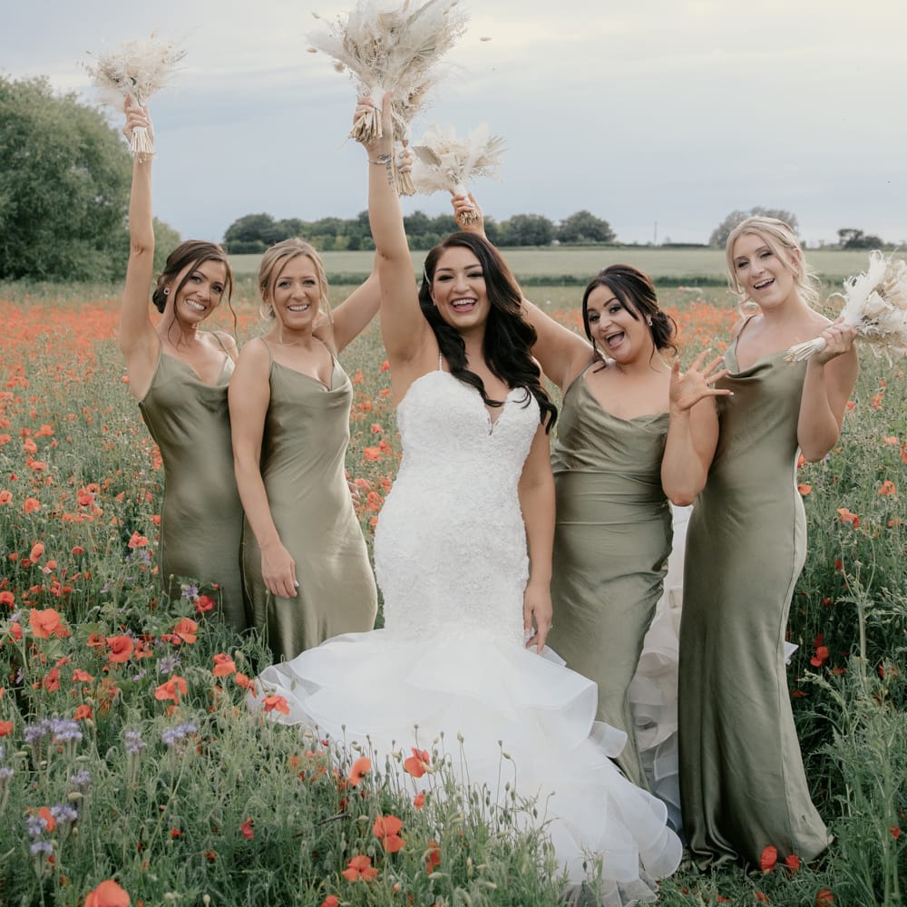 Bride in a sweetheart neckline fitted wedding dress with fishtail skirt holding her neutral dried flower bouquet in the air flanked by her bridesmaids in satin green dresses 