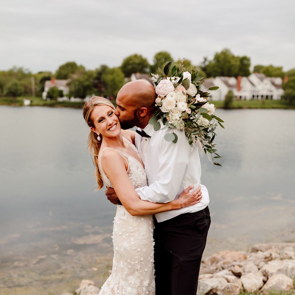 Groom in white shirt, black bow tie and white Nike Air Force 1 trainers kisses laughing bride wearing white Made With Love wedding dress on the cheek as she holds white, blush pink and green rose bridal bouquet lakeside at backyard wedding