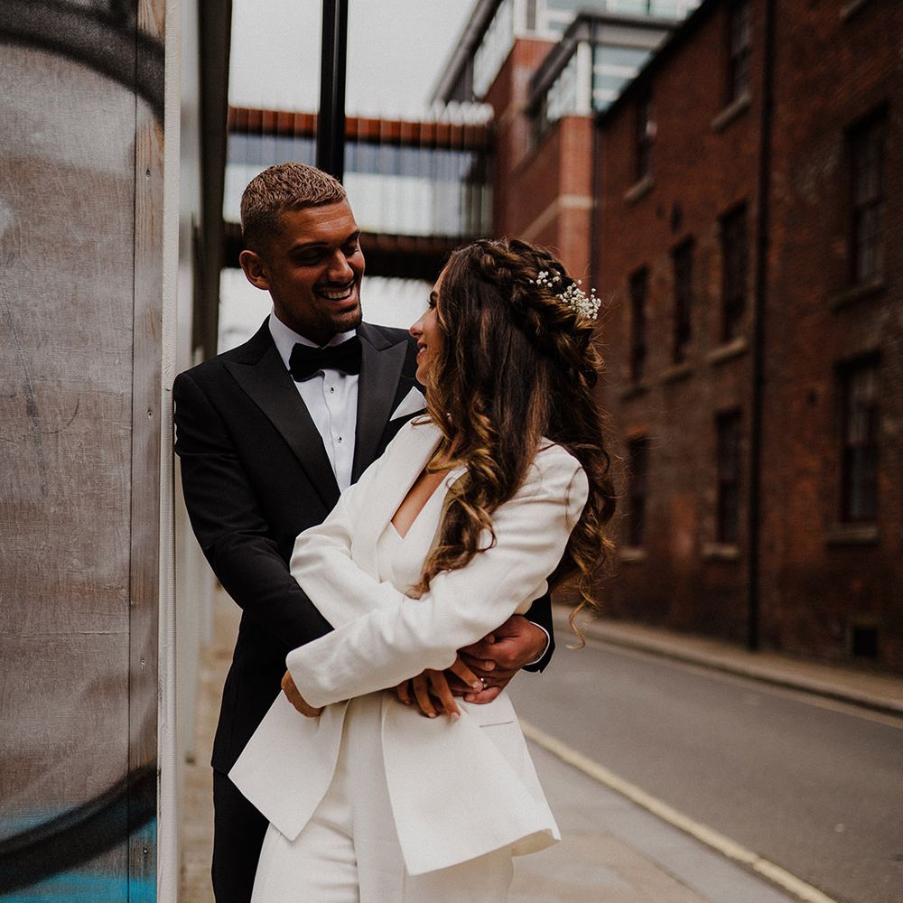 Groom in a black tuxedo embracing his bride in a white suit on the streets in Sheffield 