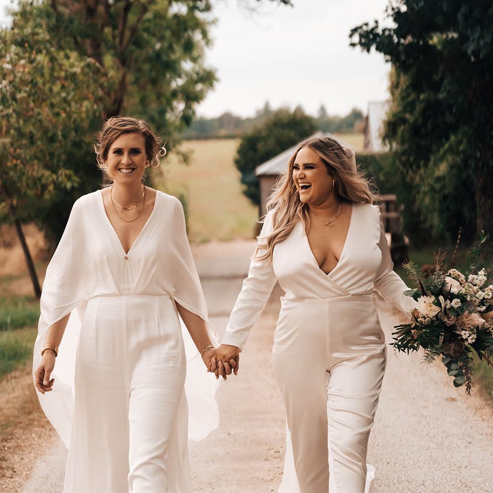 Brides in jumpsuits walk together down country lane hand in hand on their wedding day