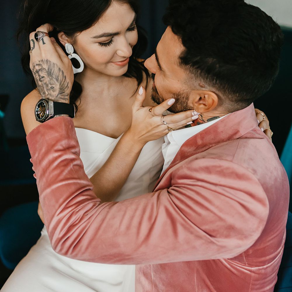 Groom in a dusky pink velvet jacket brushing his brides hair behind her ears to show off her ceramic earrings 
