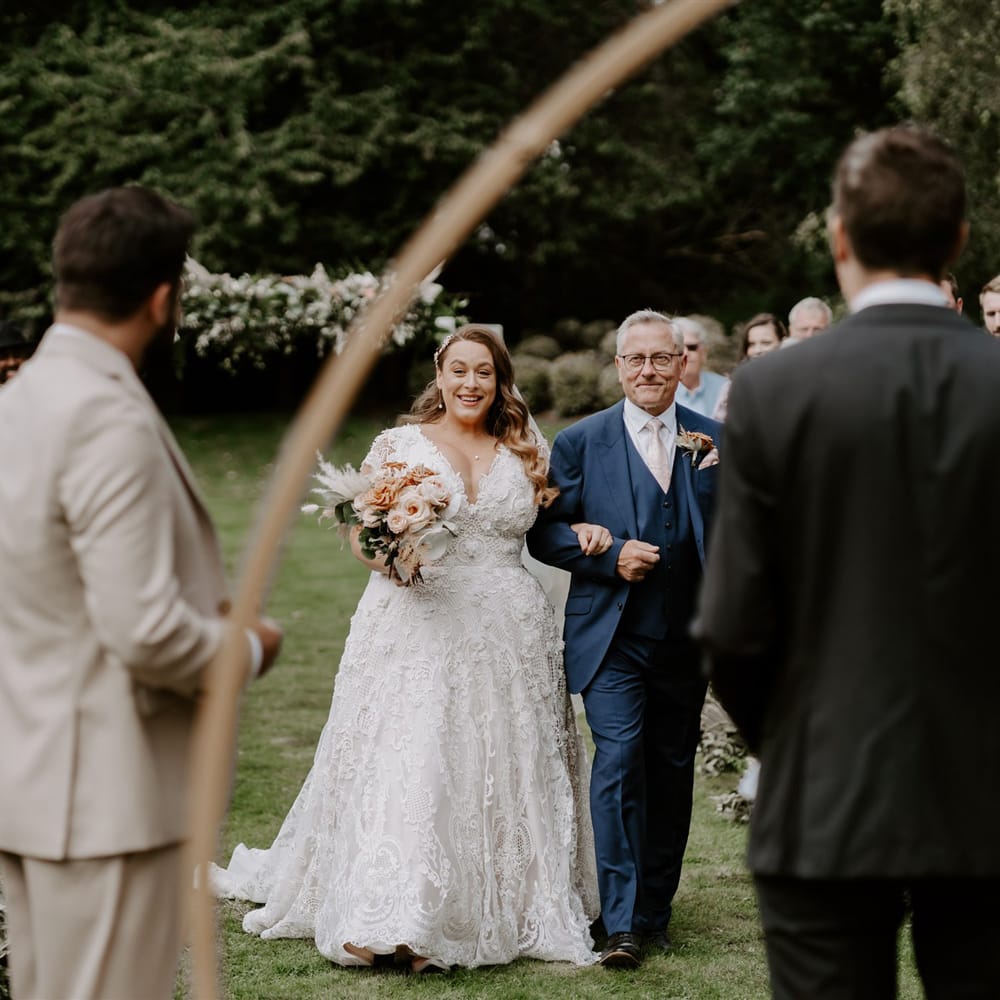Bride in homemade wedding dress holding rose bridal bouquet walks up the aisle holding arm of man in blue three piece suit during DIY garden wedding ceremony in Bedfordshire