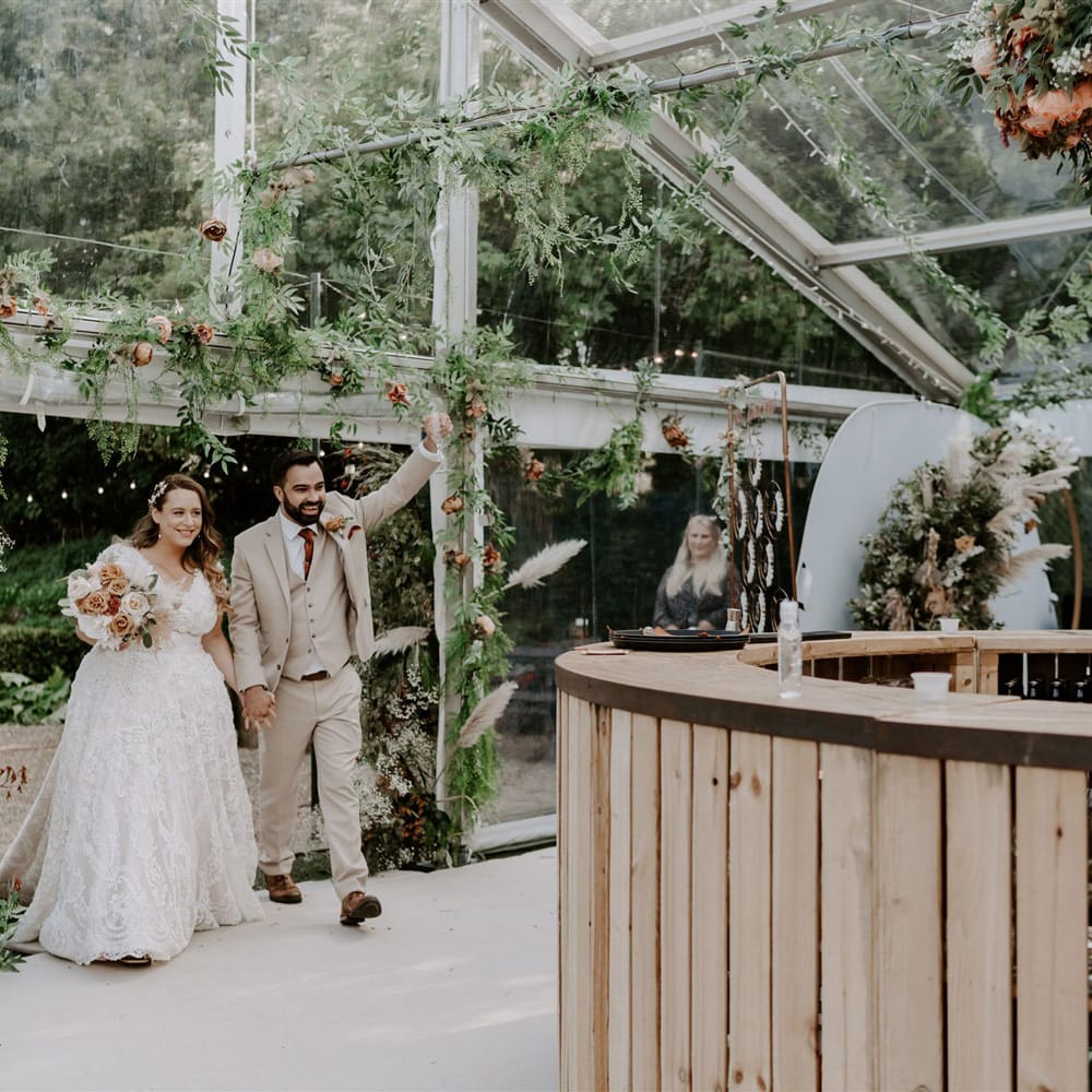 Bride in homemade lace wedding dress holding mixed rose bridal bouquet walks into marquee decorated with garlands holding hands with groom in three piece beige suit for garden wedding reception