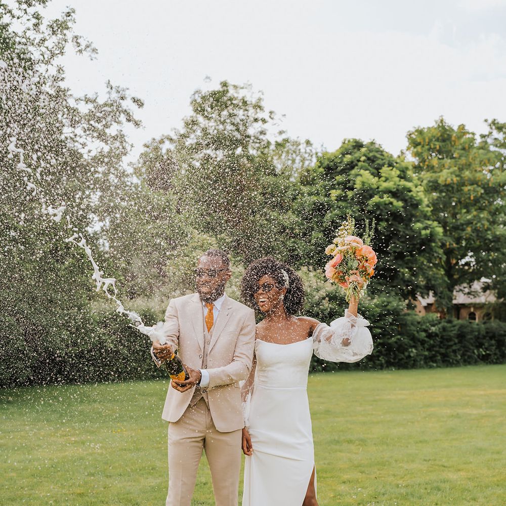 Stylish bride in a strapless wedding dress with long sheer sleeves and groom in a stone coloured suit popping champagne on the lawn 