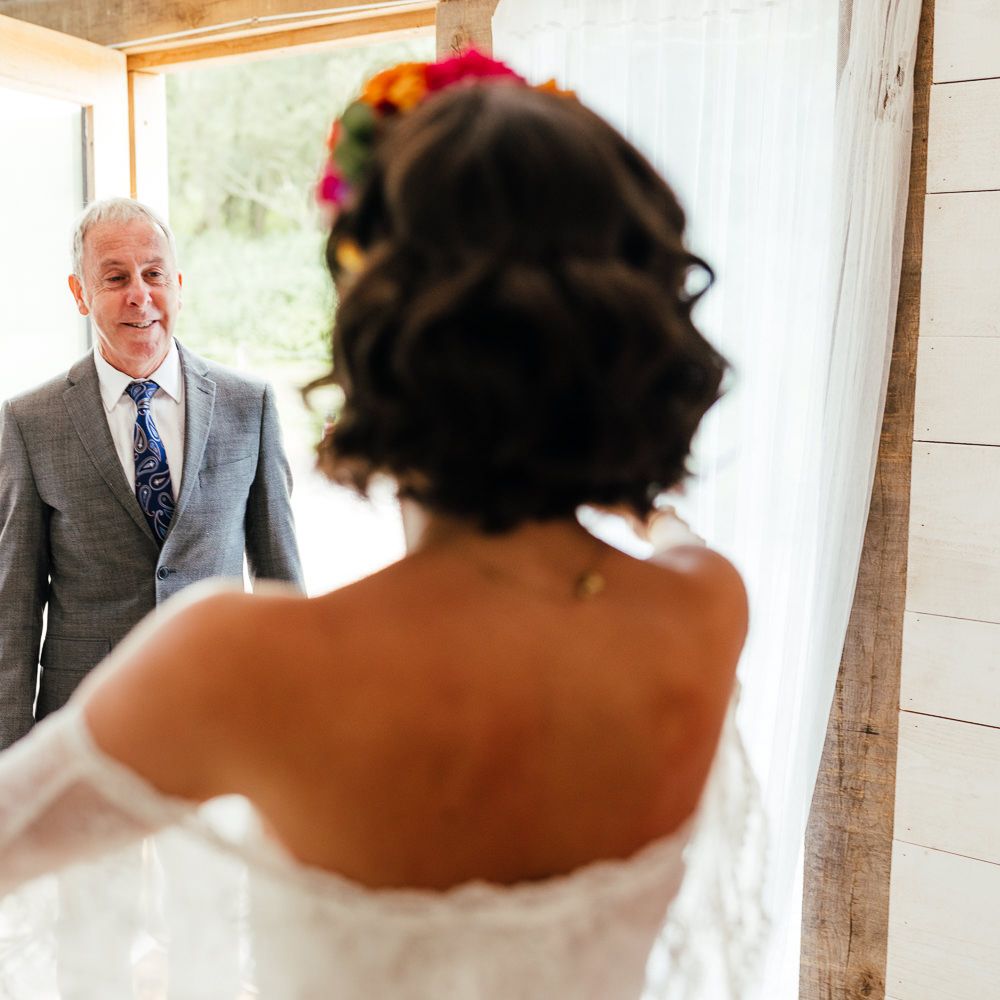 Bride in lace off the shoulder wedding dress and flower crown greens father in grey suit before outdoor wedding ceremony