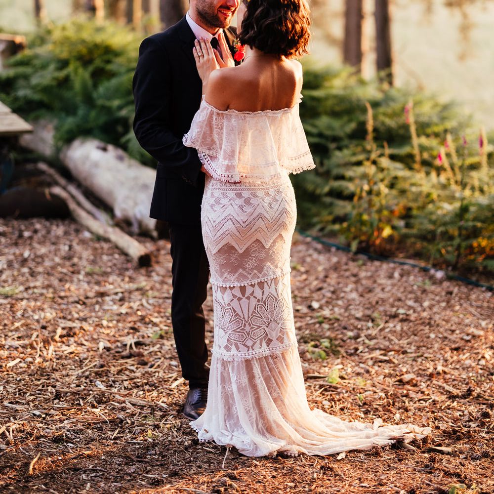 Bride in off the shoulder Grace Loves Lace wedding dress with train and colourful flower crown stands with hands on grooms chest in woodland during golden hour