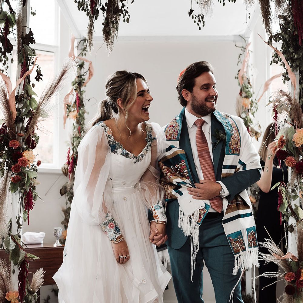 Bride in mesh sleeve embroidered Victoria Sanders wedding dress with train stands holding hands with groom in green suit, pink tie and tallit during interfaith wedding ceremony at Iscoyd Park