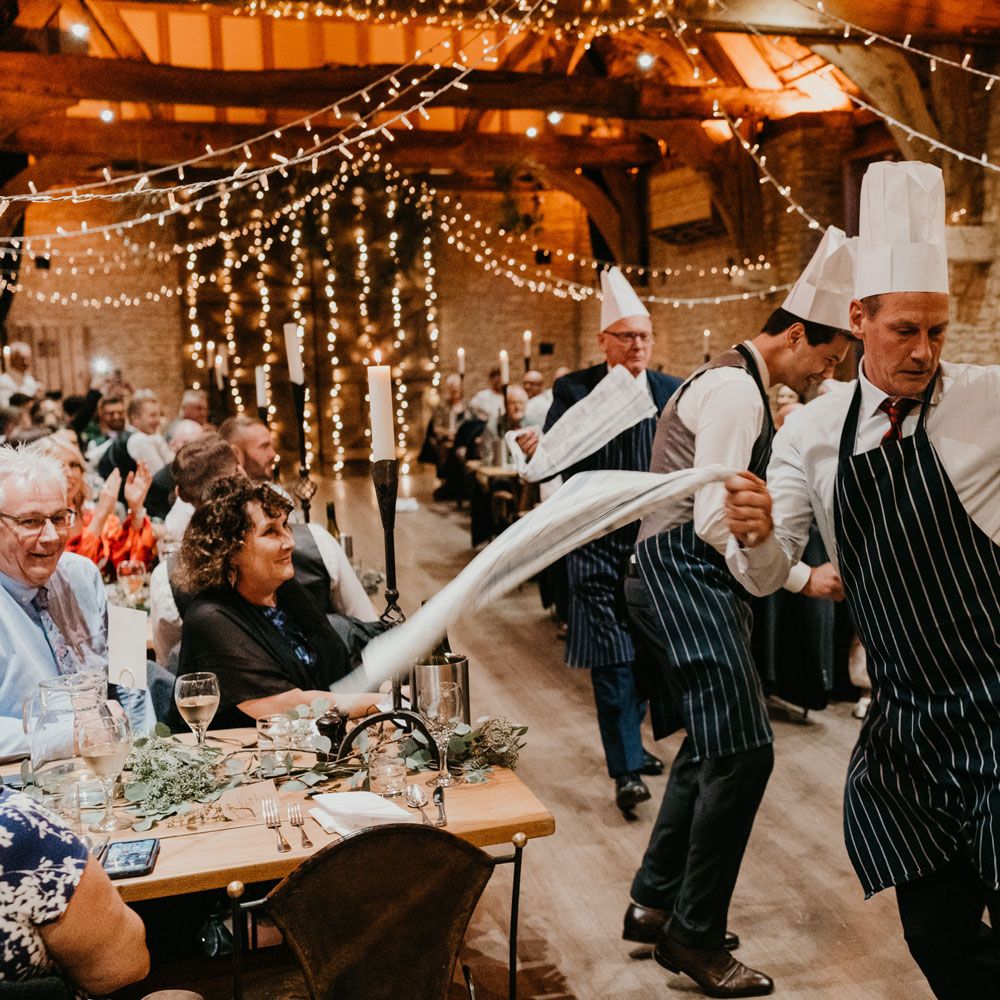Groom and guests in striped aprons and chefs hats dance with tea towels down the aisle of barn wedding breakfast 