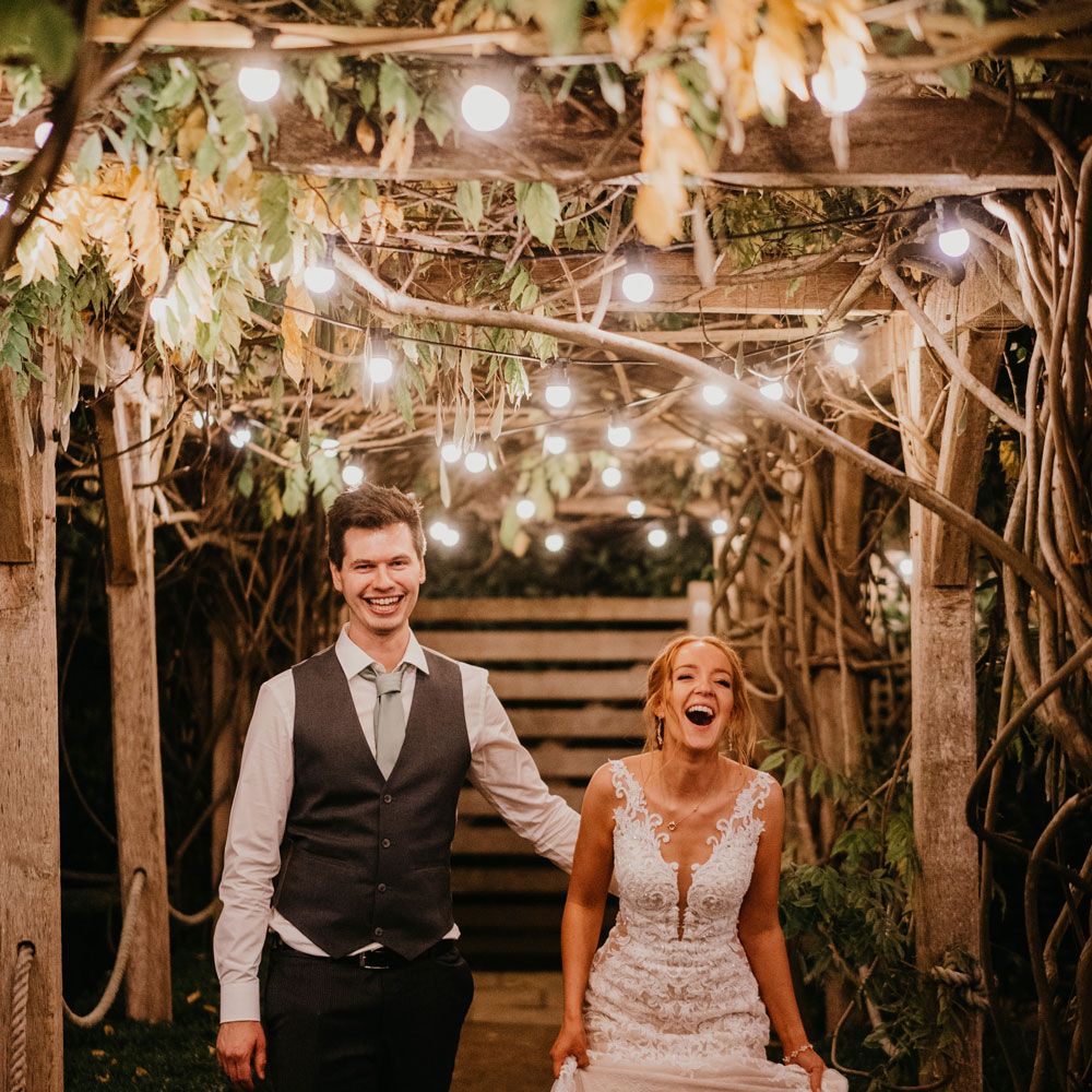 Bride in lace and tulle wedding dress stands with groom in white shirt and waistcoat on garden path lit by Edison bulbs after rustic barn wedding ceremony at The Tythe Barn in Launton