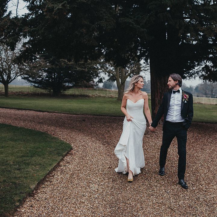 Bride holds the train of her Suzanne Neville gown as she walks with her groom who wears black tie 