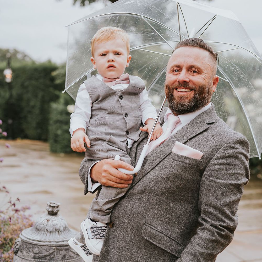 Groomsman with mini groomsman under clear umbrella on rainy day at country house wedding venue