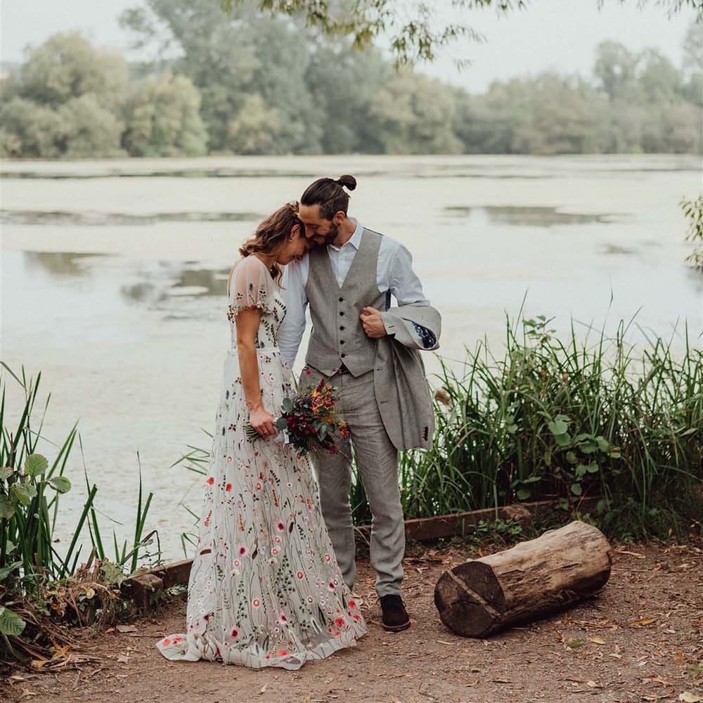 Bride in intricate floral embroidered wedding dress with groom in grey outfit stand close together at lake wedding venue 