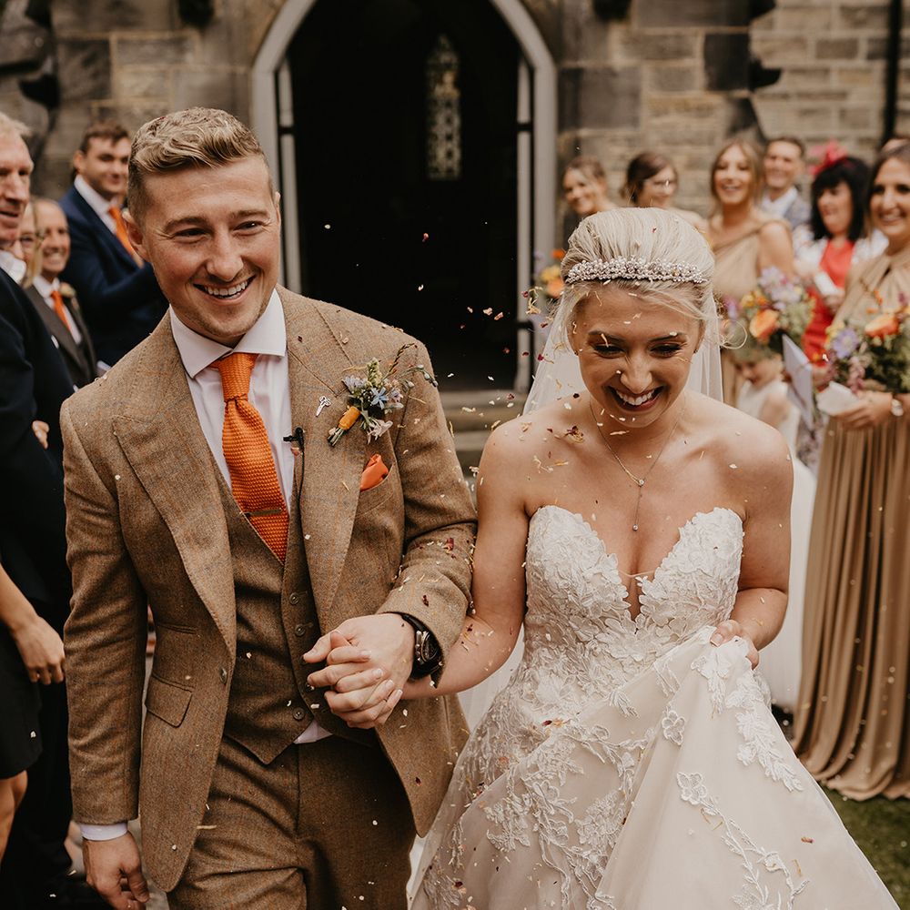 Bride in strapless sweetheart lace wedding gown with pearl tiara has confetti exit with groom in brown and orange suit and tie 