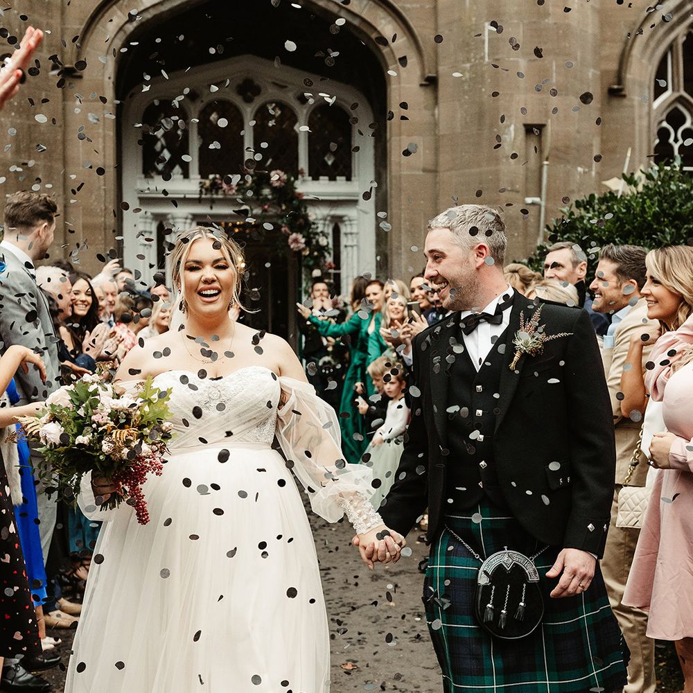 Groom smiles at the bride as she smiles at the camera as they walk through black confetti 