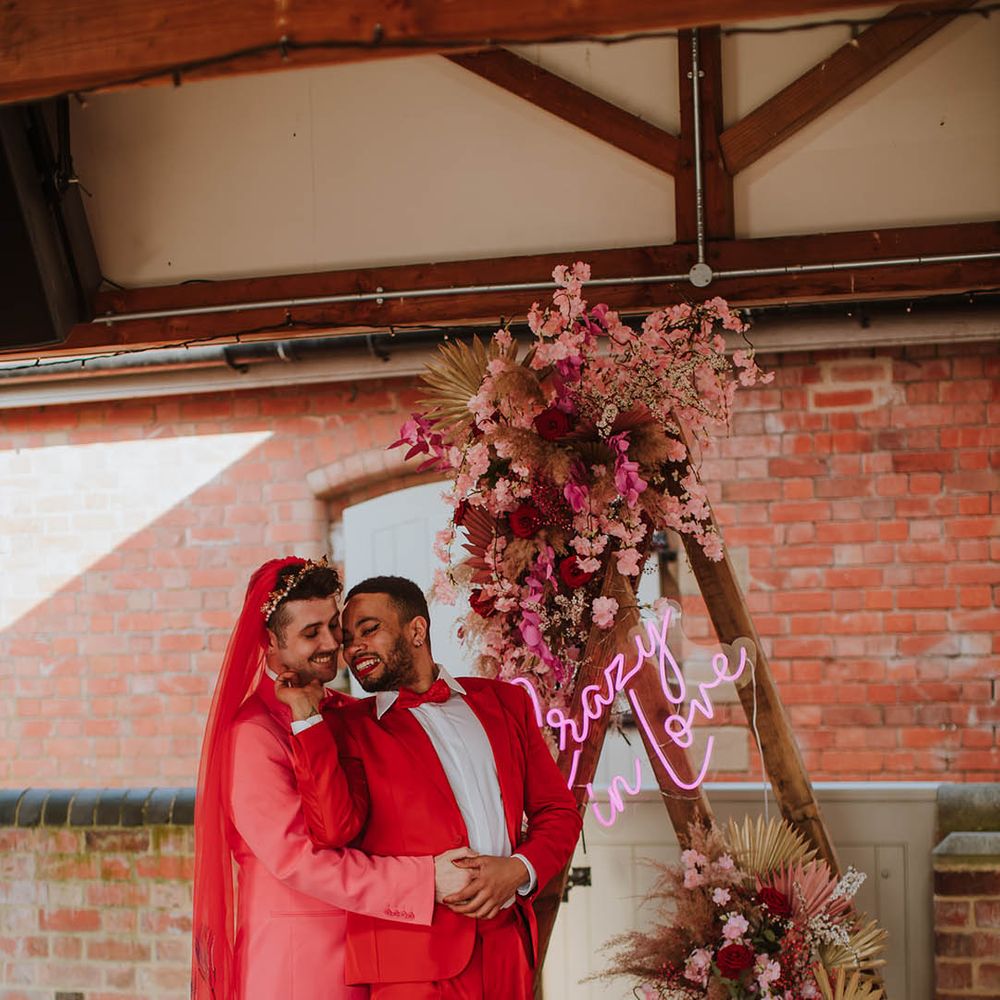 LGBTQI+ couple standing at the wooden frame altar decorated with pink flowers and dried grasses in coloured groom suits