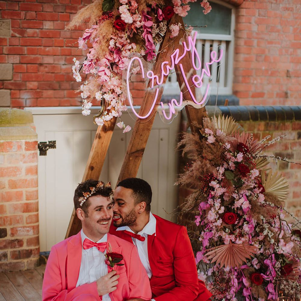 LGBTQI+ couple in pink and red suits sitting in front of a wooden triangle frame altar decorated with a pink neon sign and pink flowers and dried grass floral arrangement 
