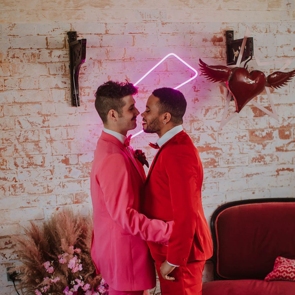 LGBTQI+ couple in pink and red suits standing in front of a pink neon sign at Prestwold Hall Barn wedding venue 