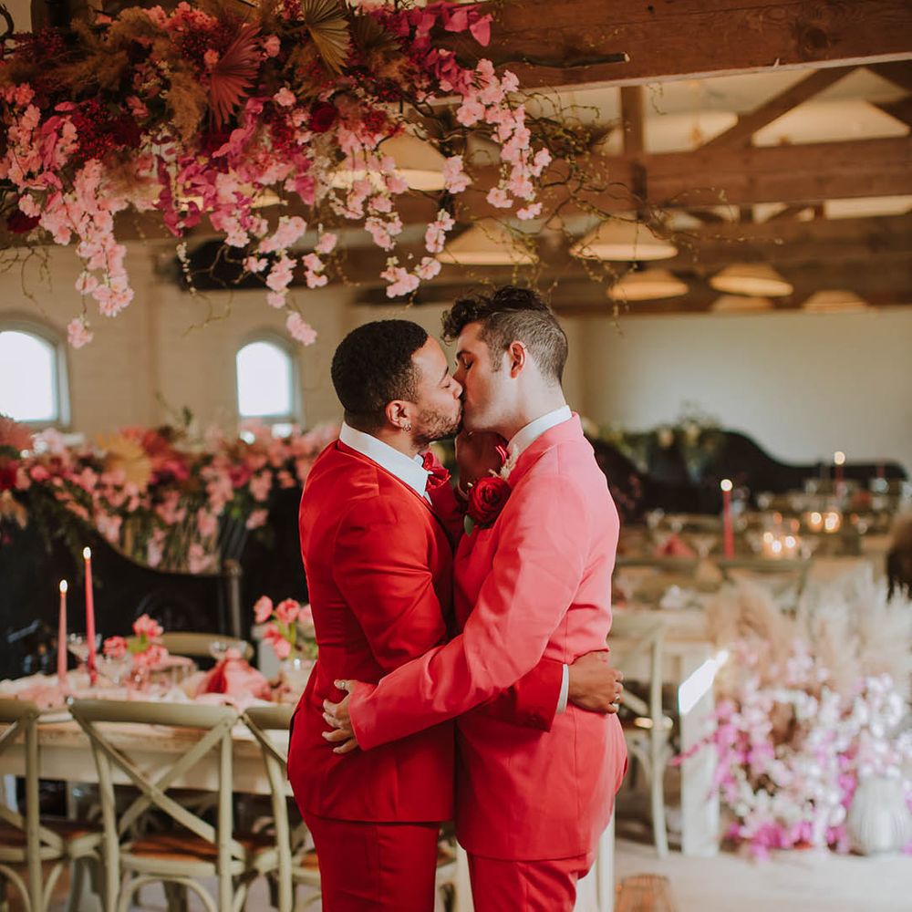 Two grooms in pink and red suits kissing at their Prestwold Hall Barn reception decorated with pink cherry blossom 