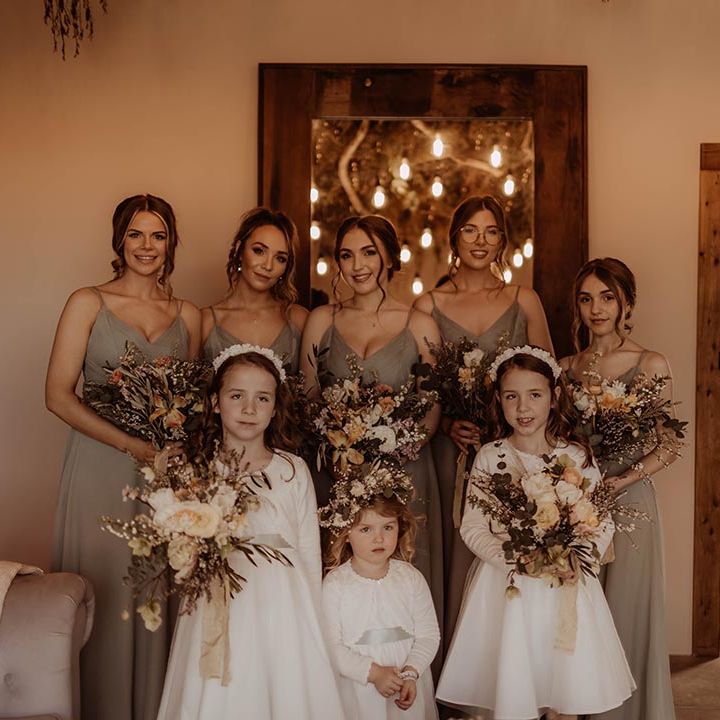 Bridesmaids holding autumnal coloured bouquets with flower girls in white dresses with cardigans and headbands or flower crowns