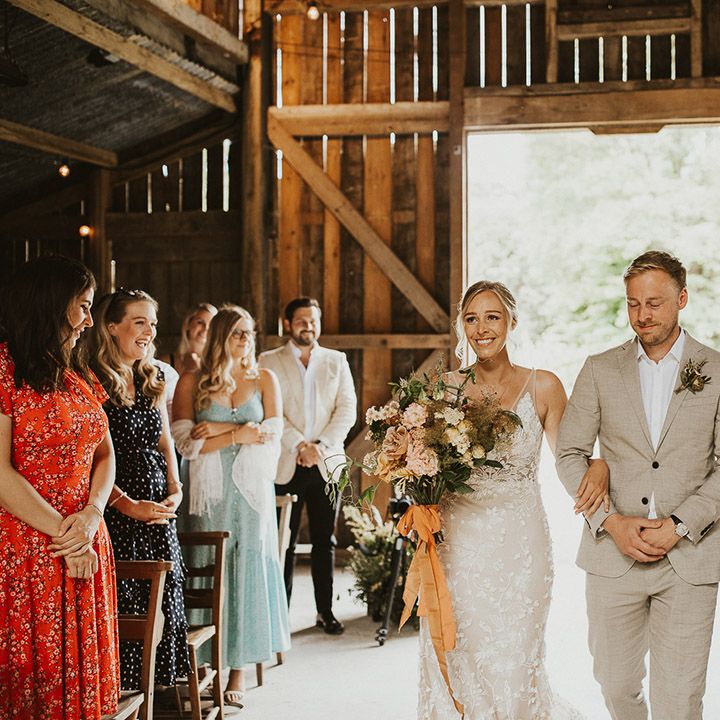 Bride smiles in a Made With Love wedding dress carrying her bouquet tied with orange ribbon as she is walked down the aisle 