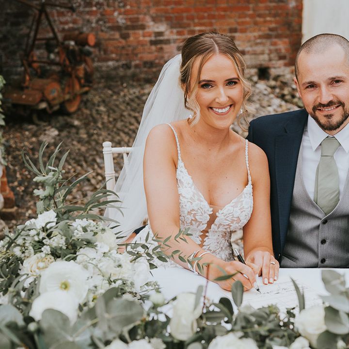 Bride in a floral lace wedding dress sitting with the groom in a dark blue suit and grey waistcoat signing the wedding register
