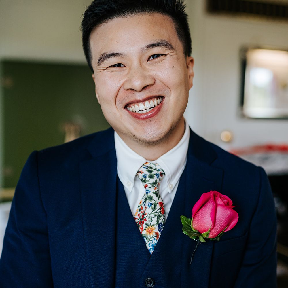 Groom wears three piece blue suit complete with embroidered floral tie and pink rose buttonhole 